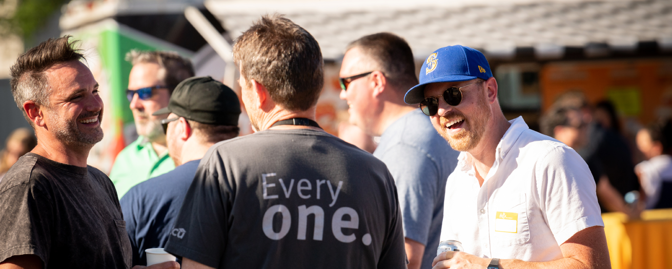 Three men, one in an STCU shirt, holding drinks and talking at an outdoor party