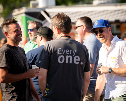 Three men, one in an STCU shirt, holding drinks and talking at an outdoor party