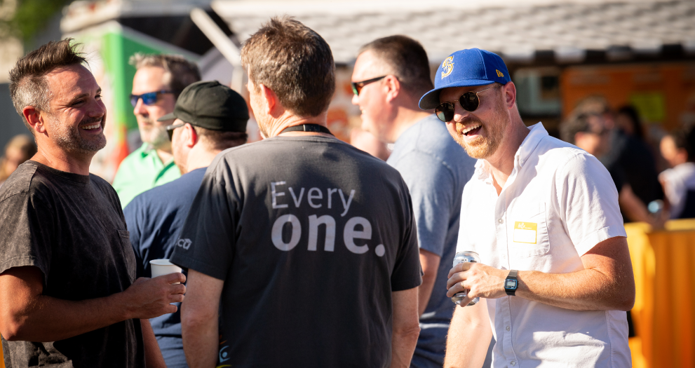 Three men, one in an STCU shirt, holding drinks and talking at an outdoor party