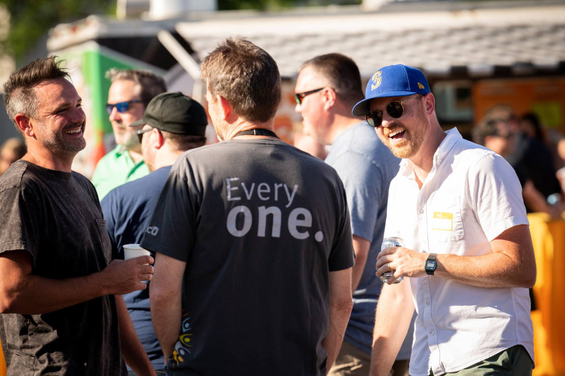 Three men, one in an STCU shirt, holding drinks and talking at an outdoor party